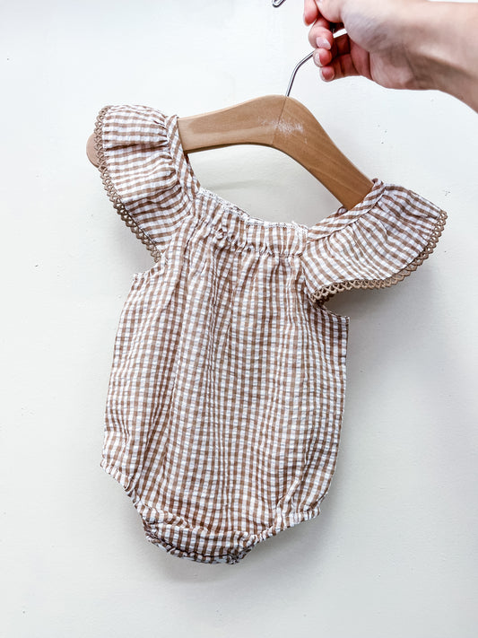A hand holds the Brown Gingham Dainty Romper by Tiny Tot Bundles—a baby romper with brown and white gingham pattern, ruffled sleeves, and decorative trim—displayed on a wooden hanger against a plain white background.