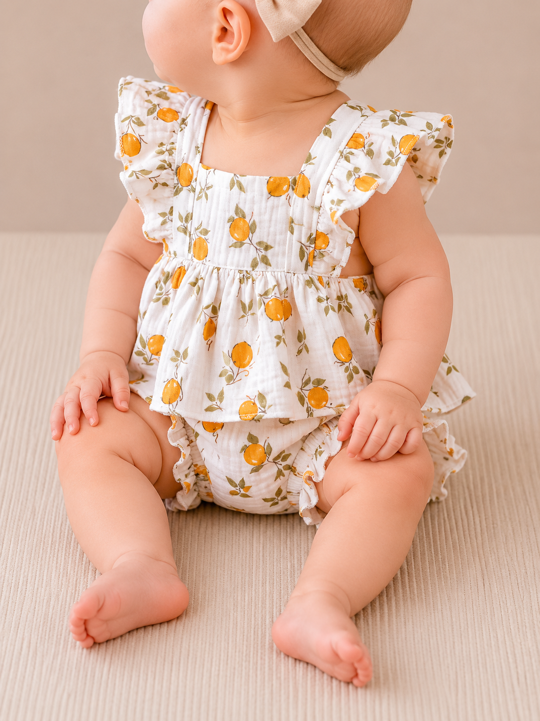 A baby models the Sunny Lemon Set by Tiny Tot Bundles—lemon-print ruffled dress and headband—sitting sideways on a beige surface, legs apart and hands on the floor.