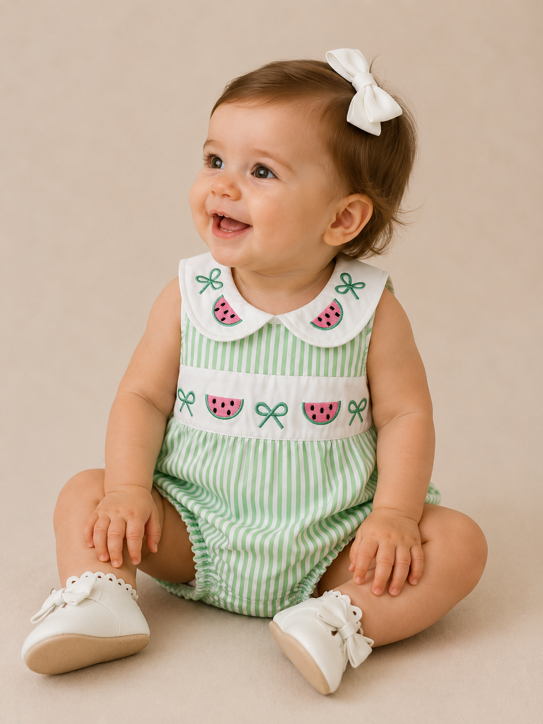A smiling baby with brown hair and a white bow sits on a beige background, wearing Tiny Tot Bundles’ Watermelon Picnic Bubble—a green and white striped romper with watermelon and bow designs—and white shoes.