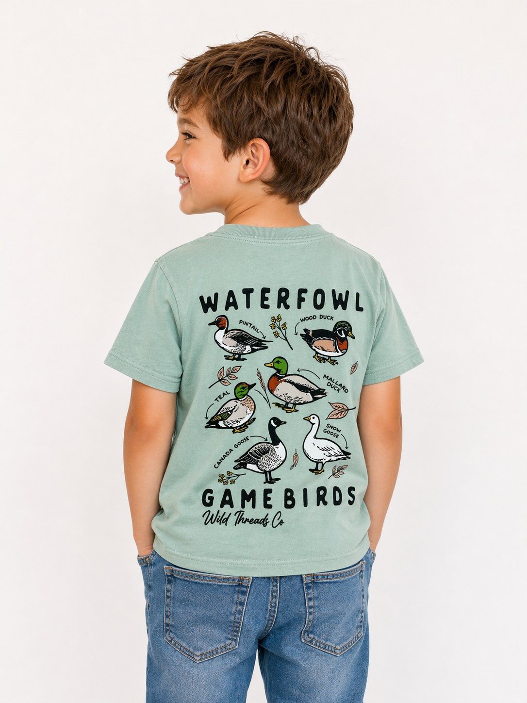 A smiling young boy wears the Little Waterfowl T-Shirt by Wild Threads Co—green with colorful illustrations and names of waterfowl and game birds on the back—paired with blue jeans, standing against a plain light background.