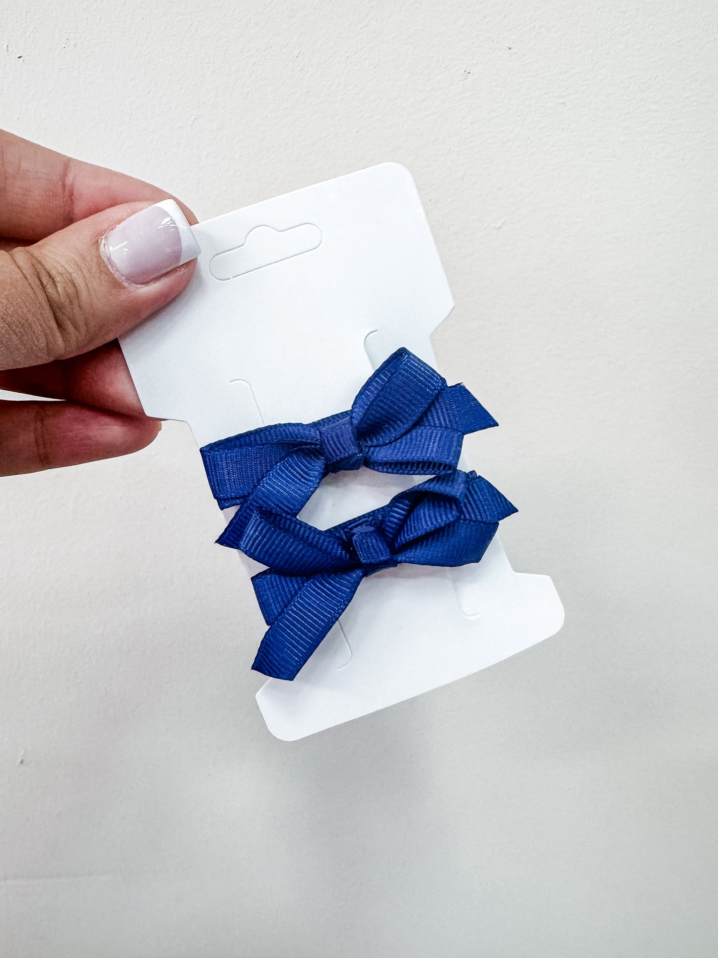 A hand with manicured nails holds a white card featuring two blue hair bows from the Duo Piggy Clip Pack by Tiny Tot Bundles, set against a plain light background.