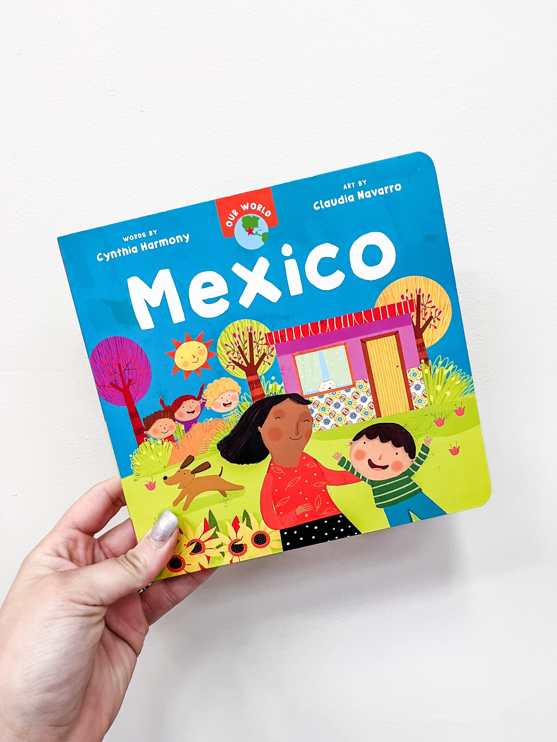 A hand holds the Barefoot Books title "Our World: Mexico," a colorful children’s book featuring illustrated kids, sunflowers, and a house on the cover.