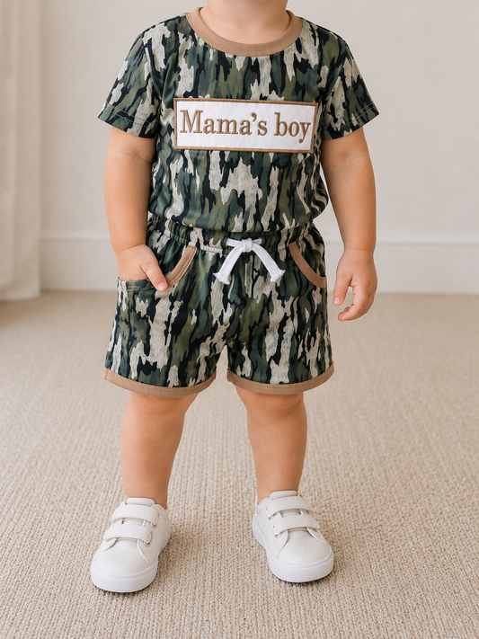A toddler models the Tiny Tot Bundles Mama’s Boy Camo Set—featuring a green camouflage outfit with "Mama’s Boy" on the shirt—and white sneakers, while standing on a beige carpet with their face not visible.