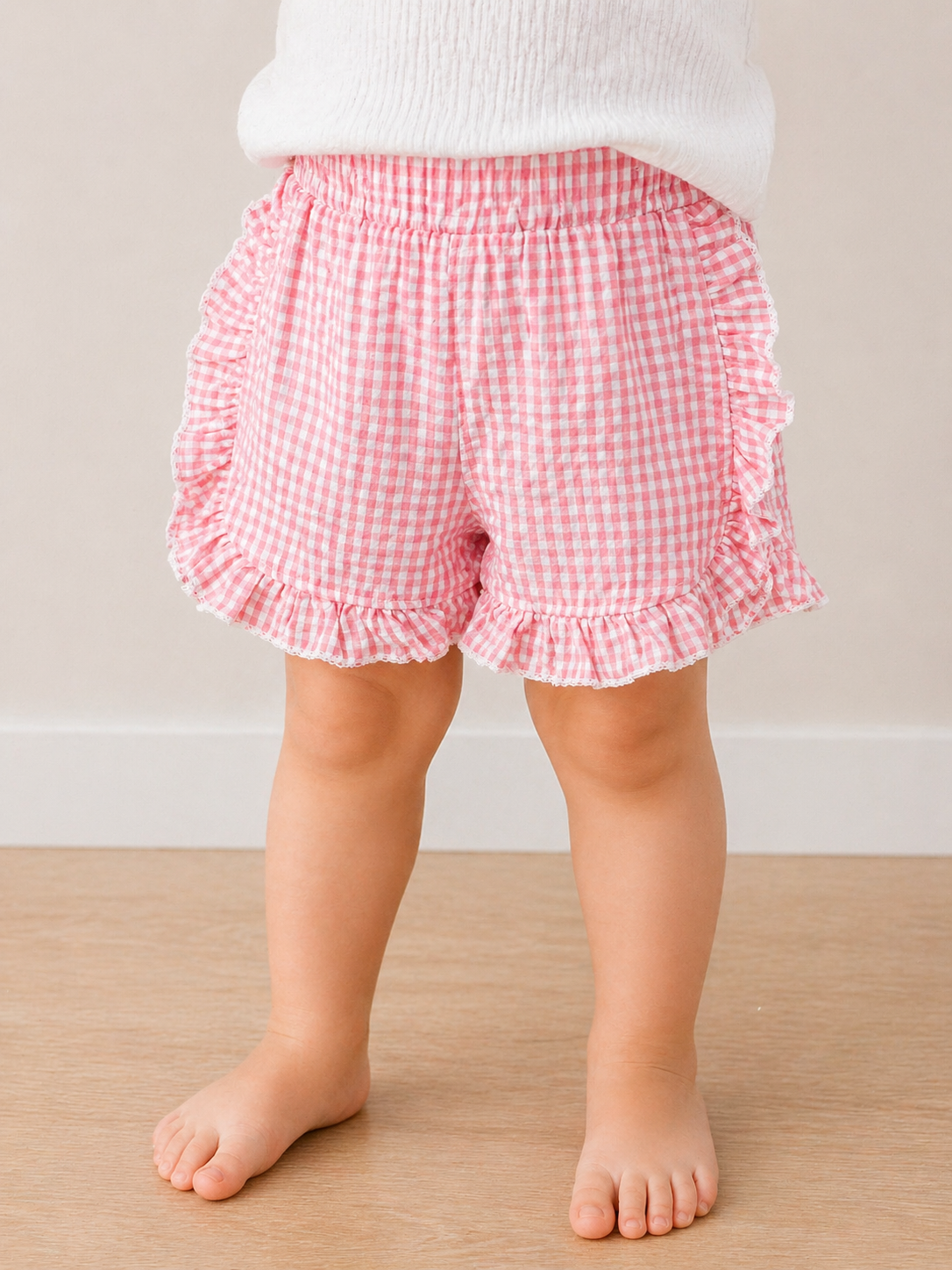 A toddler models Tiny Tot Bundles' Picnic Day Gingham Shorts—pink and white checks with ruffled edges—standing barefoot on a light wooden floor. Only the child's legs and lower torso are visible.