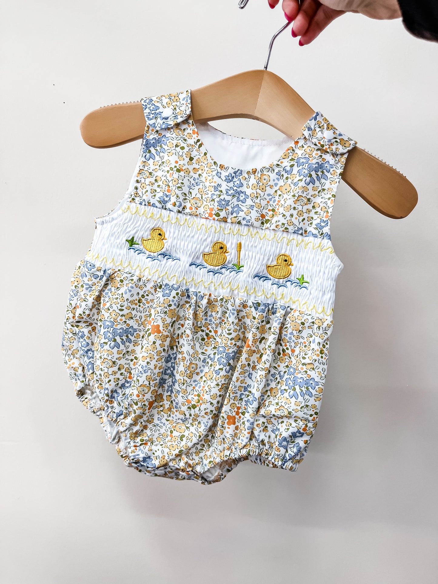 A hand holds the Tiny Tot Bundles Little Duckling Bubble—a baby romper with a yellow, blue, and white floral pattern and three embroidered yellow ducks on a smocked white chest panel.