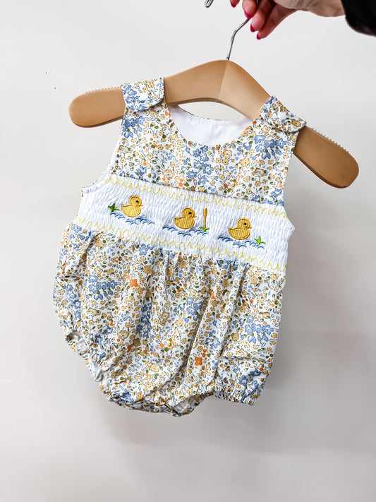 A hand holds the Tiny Tot Bundles Little Duckling Bubble—a baby romper with a yellow, blue, and white floral pattern and three embroidered yellow ducks on a smocked white chest panel.