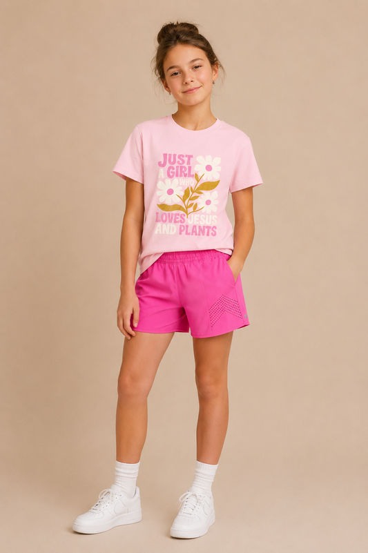 A young girl smiles in the Tiny Tot Bundles Jesus & Plants Youth Tee, light pink with "Just a girl who loves Jesus and plants" text, paired with pink shorts, white socks, and sneakers against a beige background.