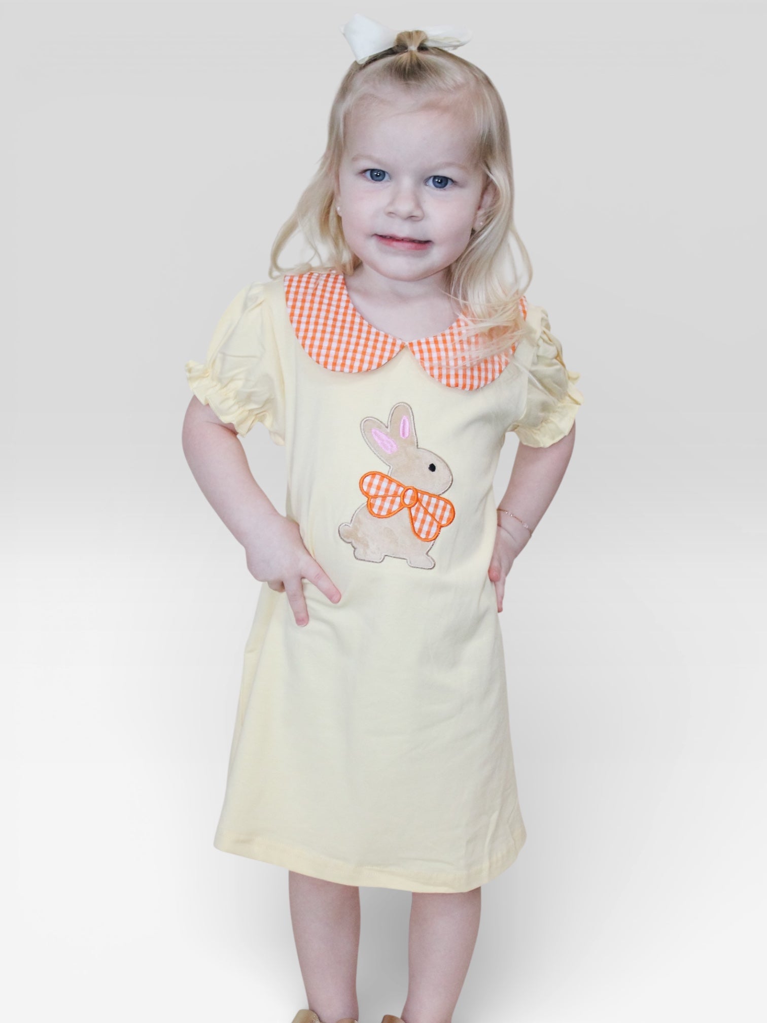 A young girl with blonde hair wears the Buttercup Bunny Dress by Tiny Tot Bundles, featuring a yellow design with an orange gingham collar and bunny applique. She smiles softly against a plain white background, hands on her hips.