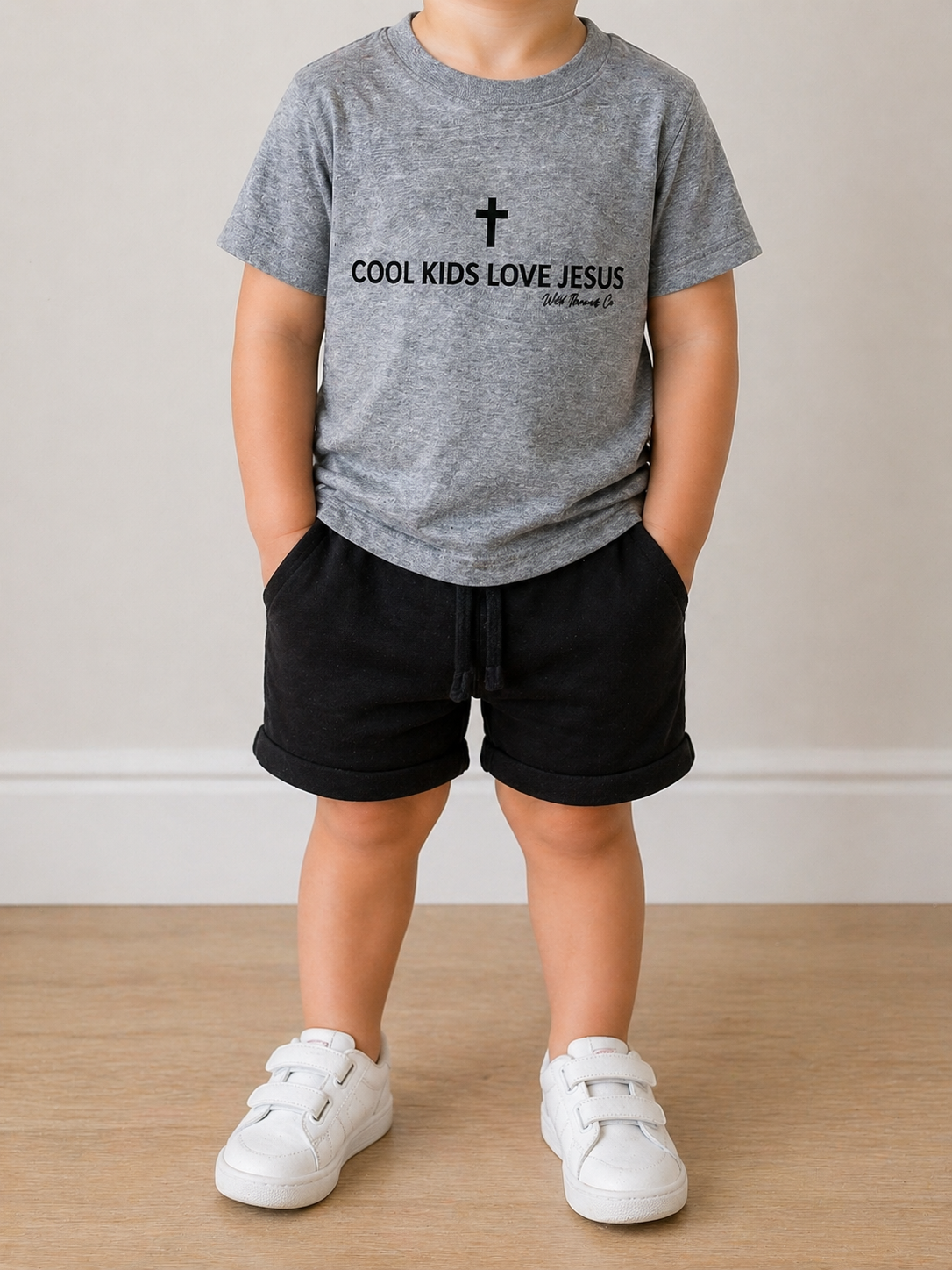 A young child stands indoors wearing the Wild Threads Co Faith Kid T-Shirt featuring "COOL KIDS LOVE JESUS" with a cross, paired with black shorts and white sneakers. Their hands are in their pockets and their face isn't visible.