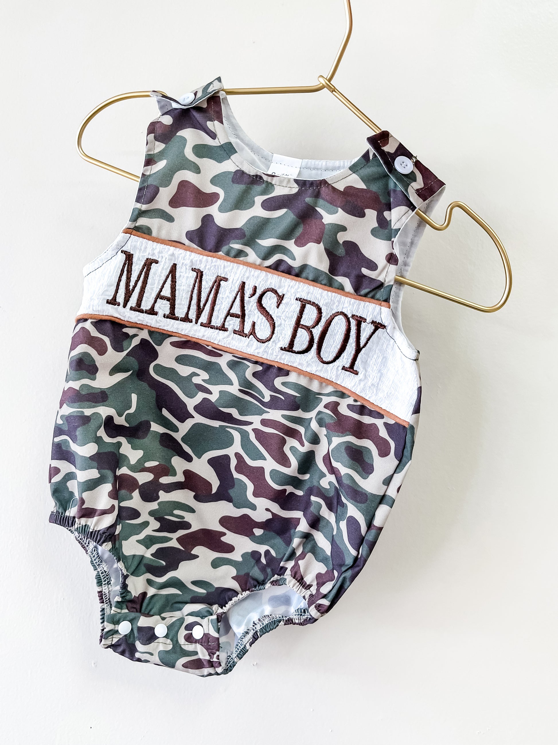 The Mama's Boy Camo Romper by Tiny Tot Bundles, in green and brown camo, hangs on a gold hanger with "MAMAS BOY" embroidered boldly across the chest.