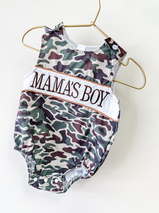 The Mama's Boy Camo Romper by Tiny Tot Bundles, in green and brown camo, hangs on a gold hanger with "MAMAS BOY" embroidered boldly across the chest.