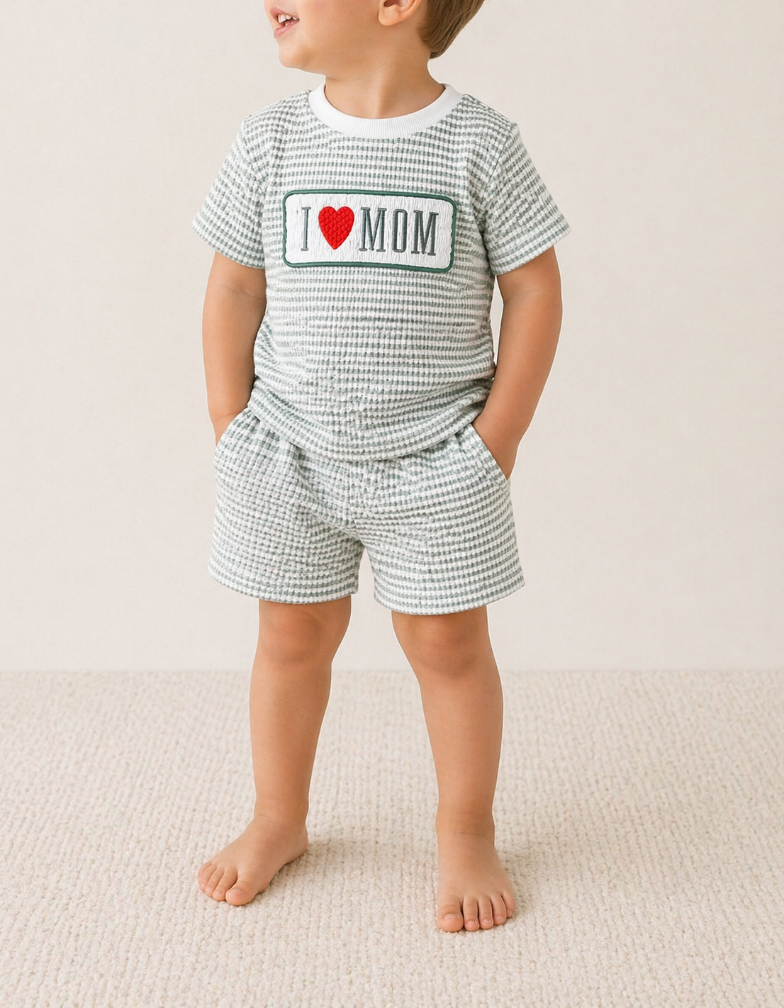 A toddler stands barefoot on a light carpet, wearing the Tiny Tot Bundles Love Mom Stripe Set—a matching light green and white checkered short-sleeve shirt with an "I ♥ MOM" patch and coordinating shorts with pockets.