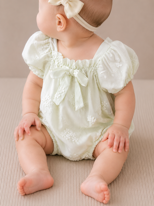 A baby in Tiny Tot Bundles' Dainty Bow Romper, a pale green outfit with puffed sleeves and bow, sits on a beige surface wearing a cream headband with a bow, looking to the side.