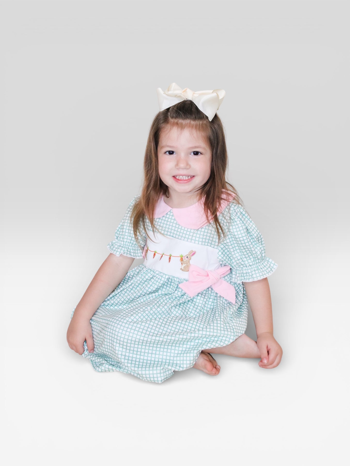 A young girl with long brown hair and a large white bow sits smiling, wearing the Tiny Tot Bundles Garden Bunny Bow Dress, a pastel checkered outfit with pink accents and an embroidered bunny, against a plain light background.