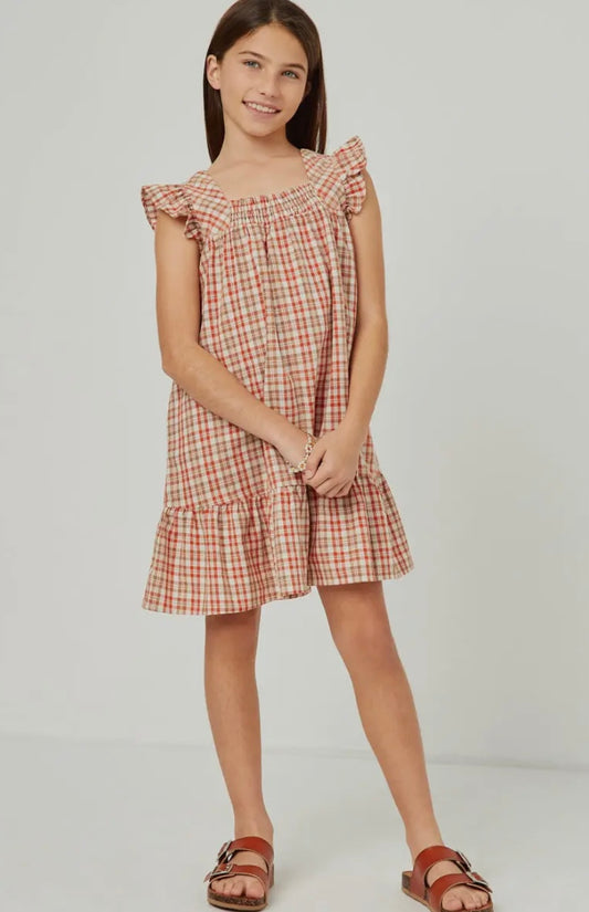 A young girl wears the Hayden LA Girls Smocked Square Neck Tartan Dress, featuring a sleeveless, ruffled design in red and beige plaid. She stands against a plain background, smiling gently with her hands clasped in front of her.