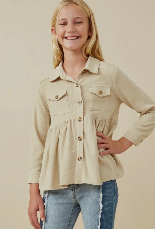 A smiling girl with long blonde hair poses in the Hayden LA Girls Linen Blend Peplum Button Up Shirt, featuring two chest pockets, paired with light blue distressed jeans against a neutral background.