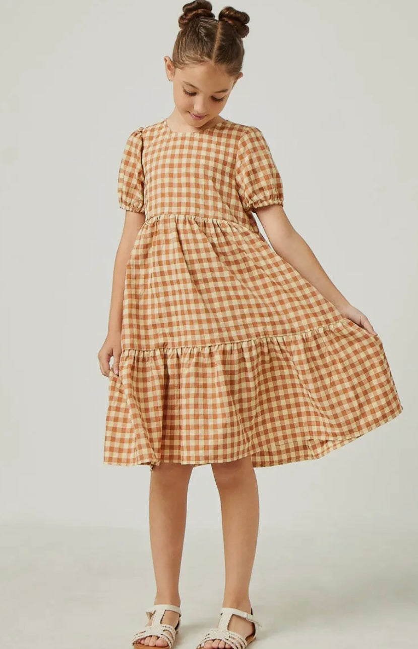 A young girl in the Hayden LA Girls Puff Sleeve Gingham Tiered Dress with short puff sleeves and a ruffled hem stands against a plain background, holding out one side of her dress and wearing white sandals.