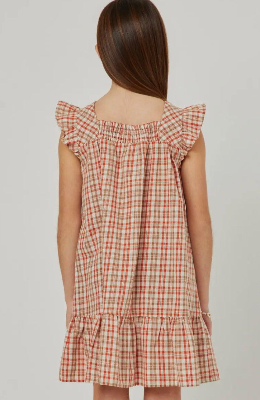 A young girl with straight brown hair faces away, wearing the Hayden LA Girls Smocked Square Neck Tartan Dress, featuring a red and beige plaid pattern and ruffled hem, against a plain background.
