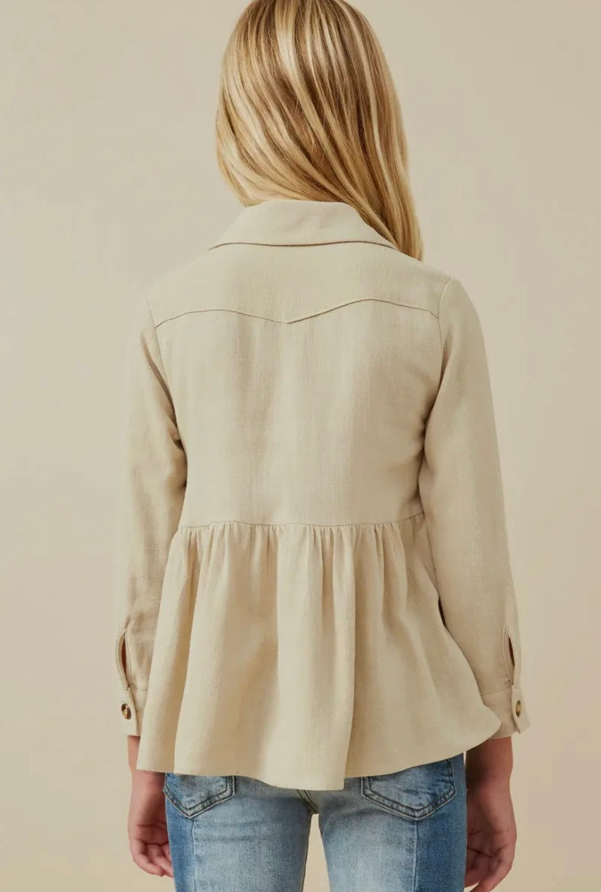 A young person with long blonde hair is seen from the back, wearing a beige Hayden LA Girls Linen Blend Peplum Button Up Shirt and blue jeans against a plain, light background.