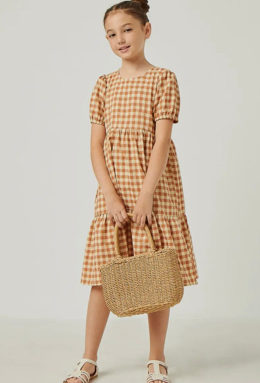 A young girl wears the Hayden LA Girls Puff Sleeve Gingham Tiered Dress, featuring brown-and-white checks and short puff sleeves, paired with a woven straw handbag, white sandals, and her hair in a bun.