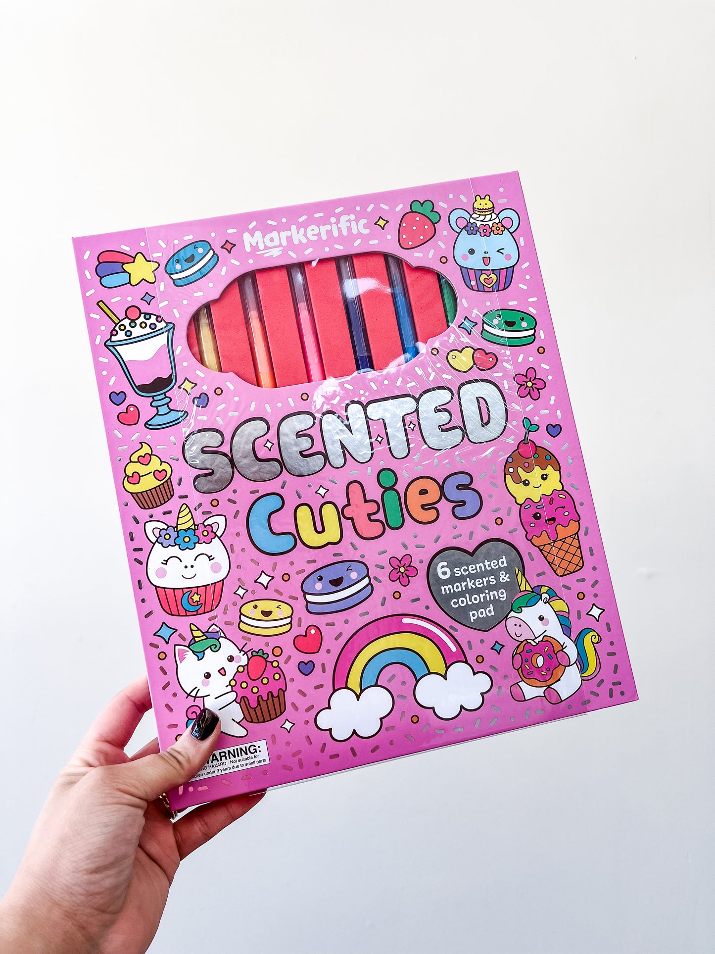 A hand holds the Tiny Tot Bundles Markerific - Scented Cuties Coloring Pad, featuring six markers in pink packaging with fun unicorn, rainbow, cupcake, and star illustrations.