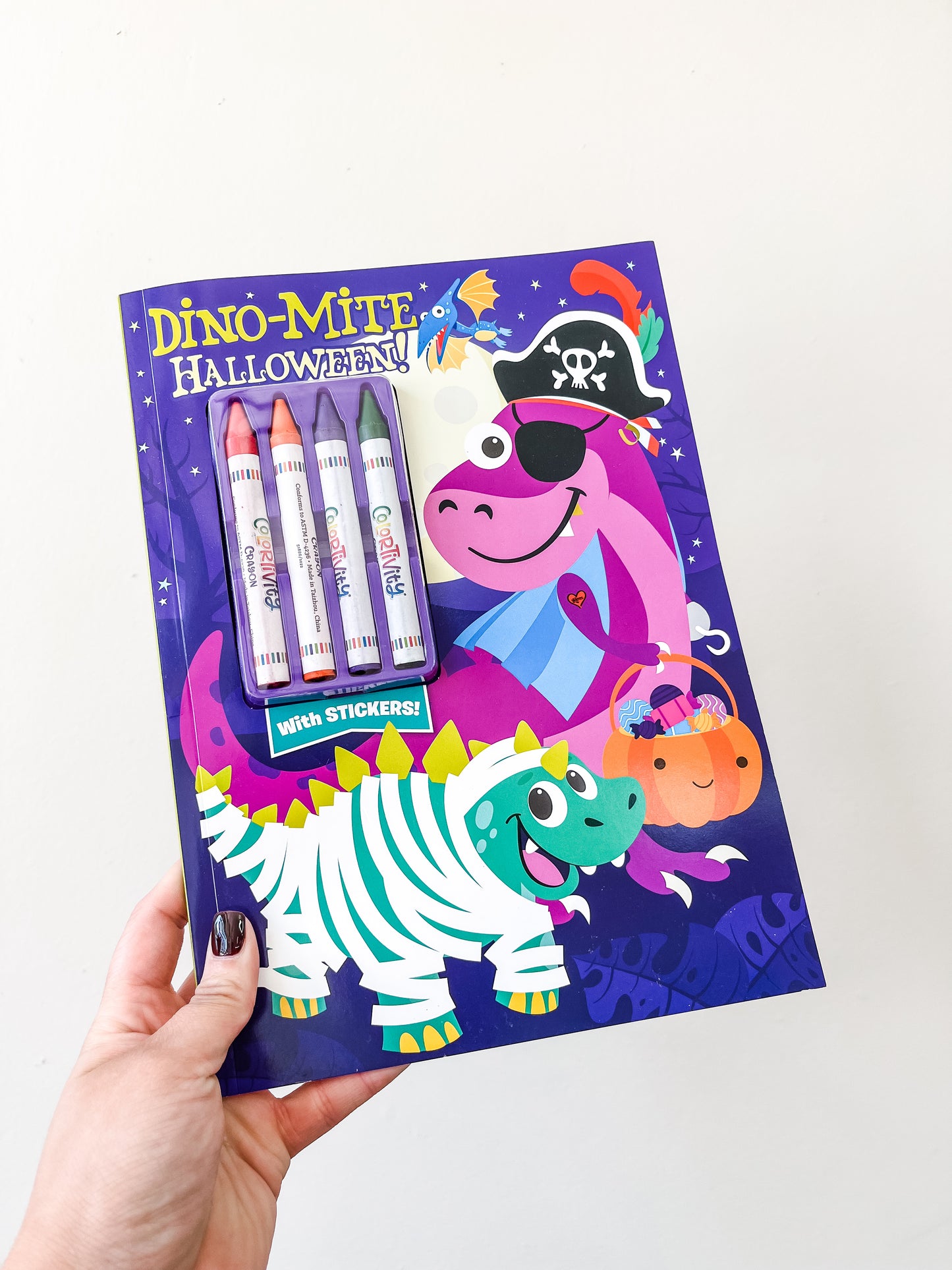 A hand holds Tiny Tot Bundles' Dino-mite Halloween Coloring Book, featuring cartoon dinosaurs in Halloween costumes, a pumpkin illustration, four markers, and a "with stickers!" label on a purple cover.