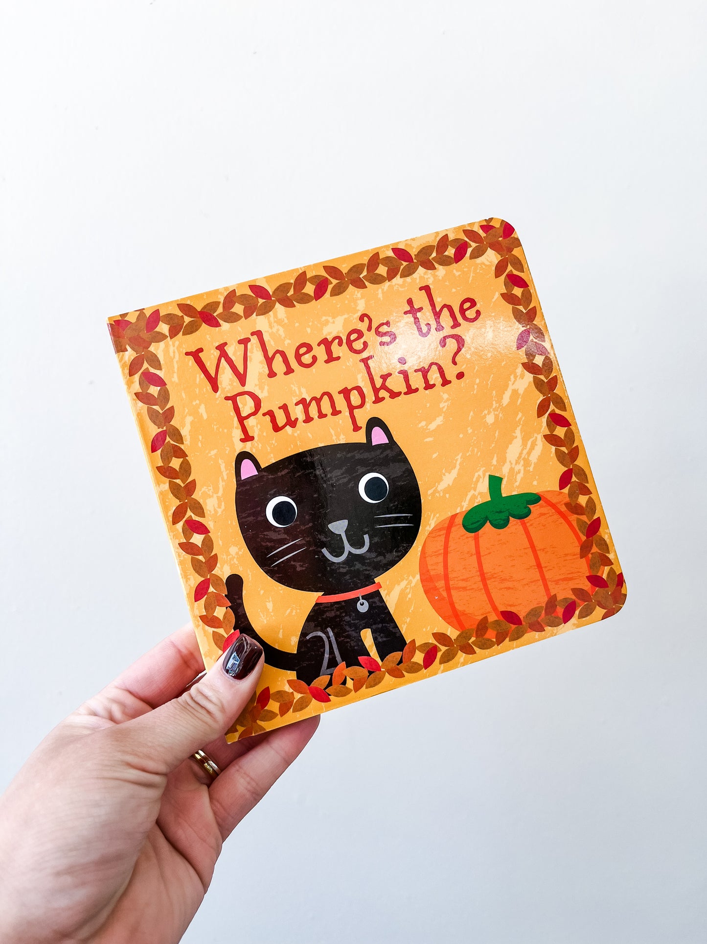 A hand holds Tiny Tot Bundles' "Where's The Pumpkin Book," featuring a cover with a black cat, orange pumpkin, and autumn leaves on a yellow background.