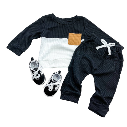 The Midnight Waffle Set by Tiny Tot Bundles includes a long-sleeve black and white top with a brown pocket, matching black pants with a white drawstring, and black baby shoes with white laces.
