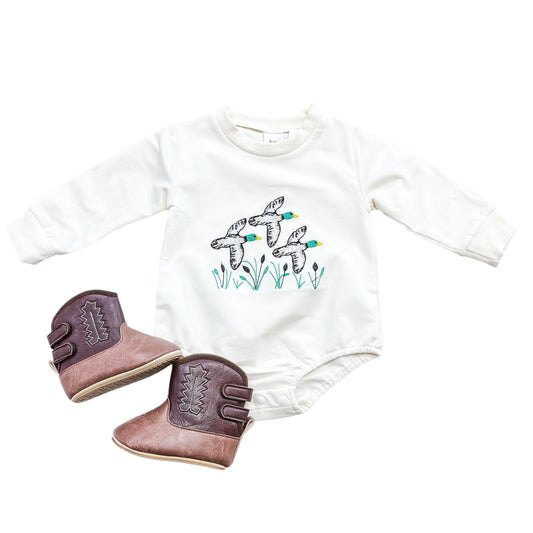 The Mallard Meadow Bubble by Tiny Tot Bundles is a white baby onesie with three birds and green grass, styled with small brown cowboy boots featuring decorative stitching.