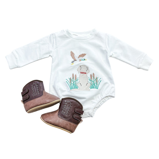 The Meadow Pup Bubble by Tiny Tot Bundles, a white baby onesie with an embroidered duck and flying birds, is paired with brown and tan cowboy boots and displayed on a white background.