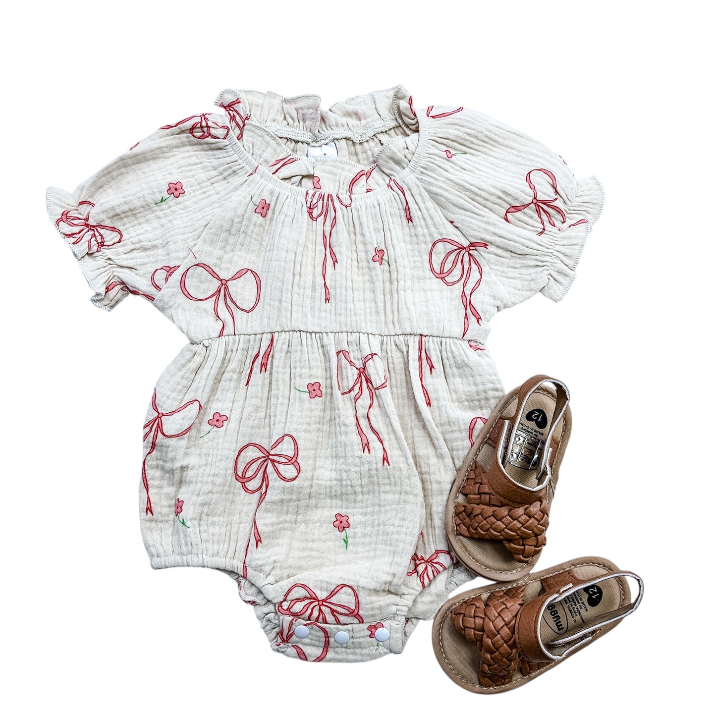 The Sweet Ribbon Bubble by Tiny Tot Bundles, a cream-colored baby romper with red bows and flower prints, is styled with brown woven sandals and displayed on a white background.