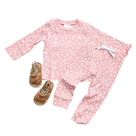 The Blush Daisy Ribbed Set by Tiny Tot Bundles, featuring a pink long-sleeve top with white polka dots, matching pants with a white bow, and brown woven baby sandals, is arranged on a white background.