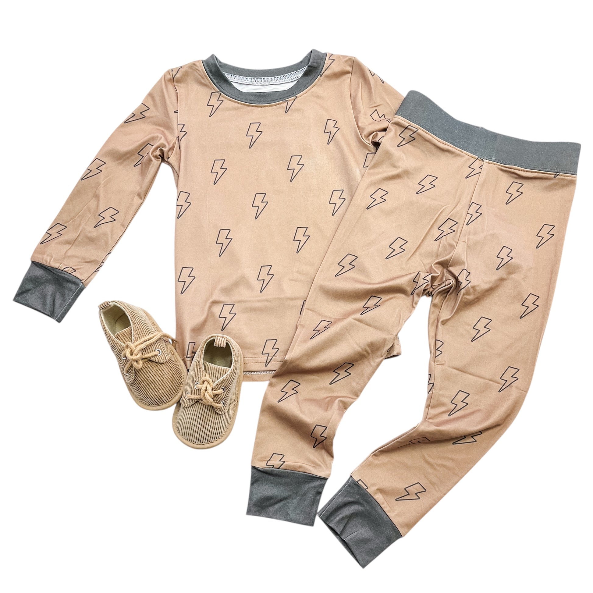 The Tiny Tot Bundles Thunderbolt Jammies Set features a two-piece tan pajama set with black lightning bolt pattern, long sleeves, gray cuffs, and matching pants. A pair of tan lace-up shoes is placed beside the pajamas.