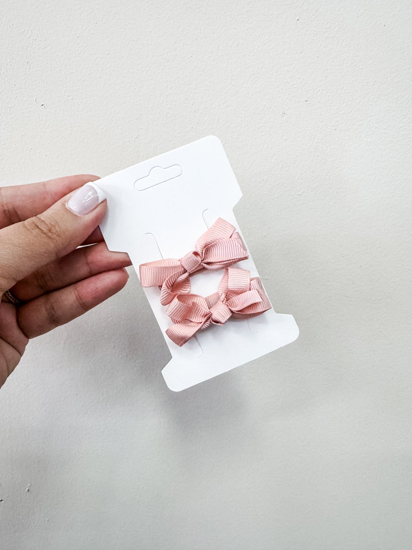 A hand with light-colored nails holds a white card featuring two small pink bows from the Tiny Tot Bundles Duo Piggy Clip Pack, set against a plain light background.