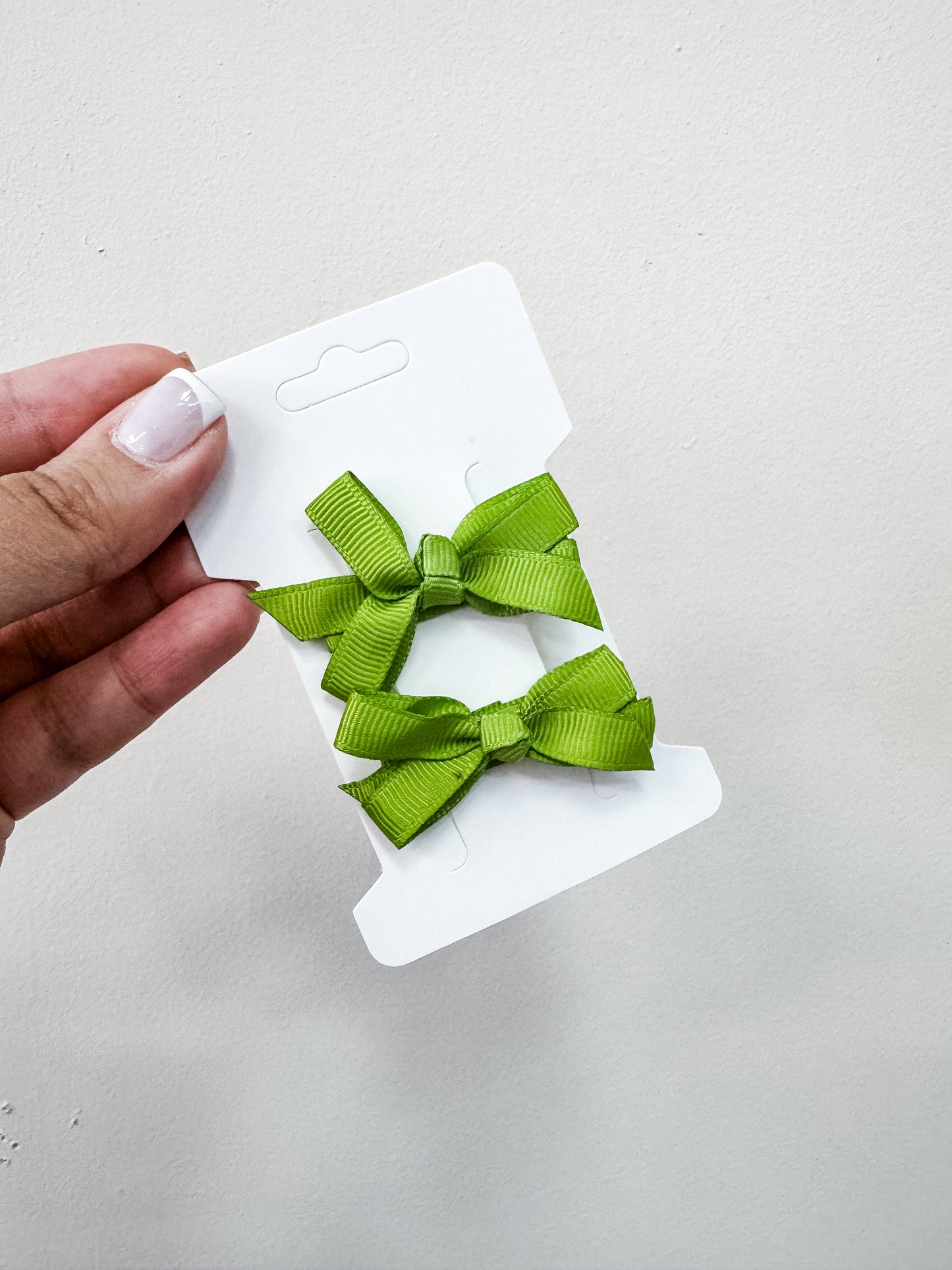 A hand with manicured nails holds a white card displaying the Duo Piggy Clip Pack by Tiny Tot Bundles—two small, neatly tied green ribbon bows—against a plain light background.