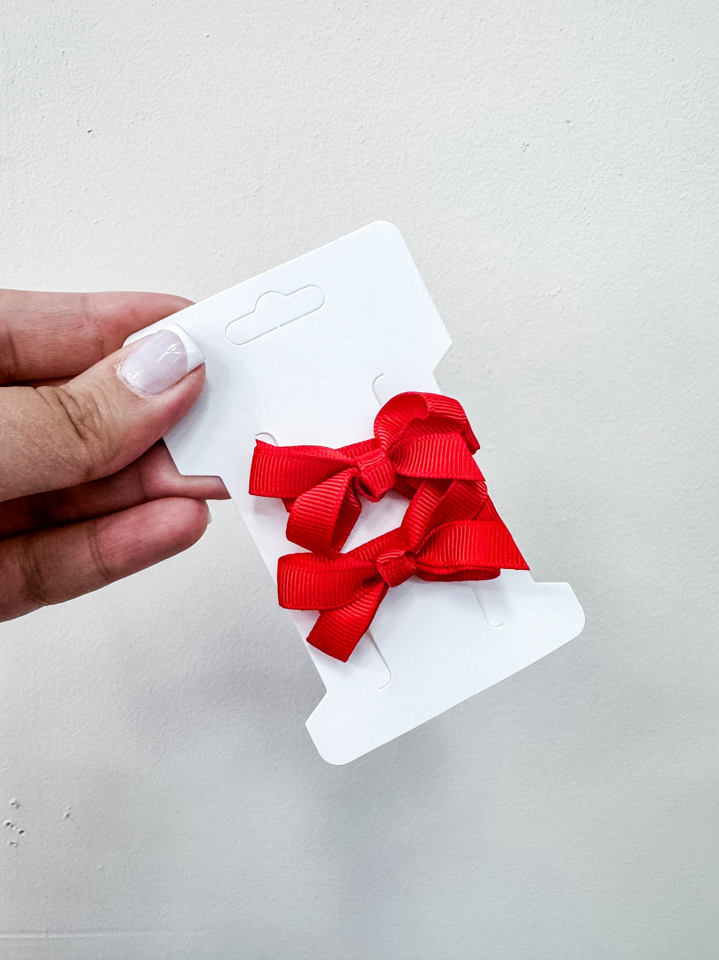 A hand holds a white card displaying two bright red bows from the Duo Piggy Clip Pack by Tiny Tot Bundles, set against a plain light background.