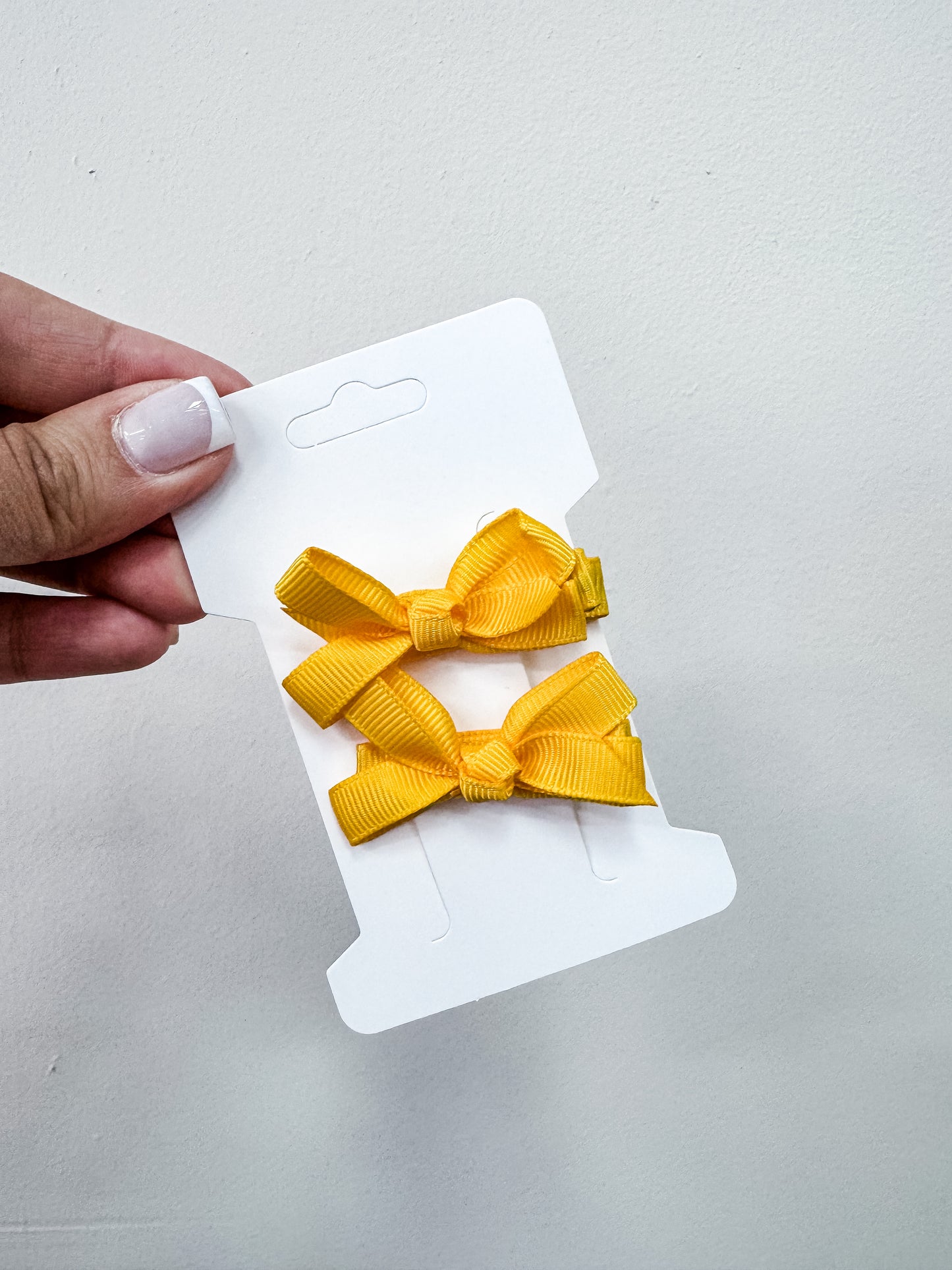 A hand with manicured nails holds a white card featuring two yellow ribbon hair bows from the Tiny Tot Bundles Duo Piggy Clip Pack, set against a plain light background.