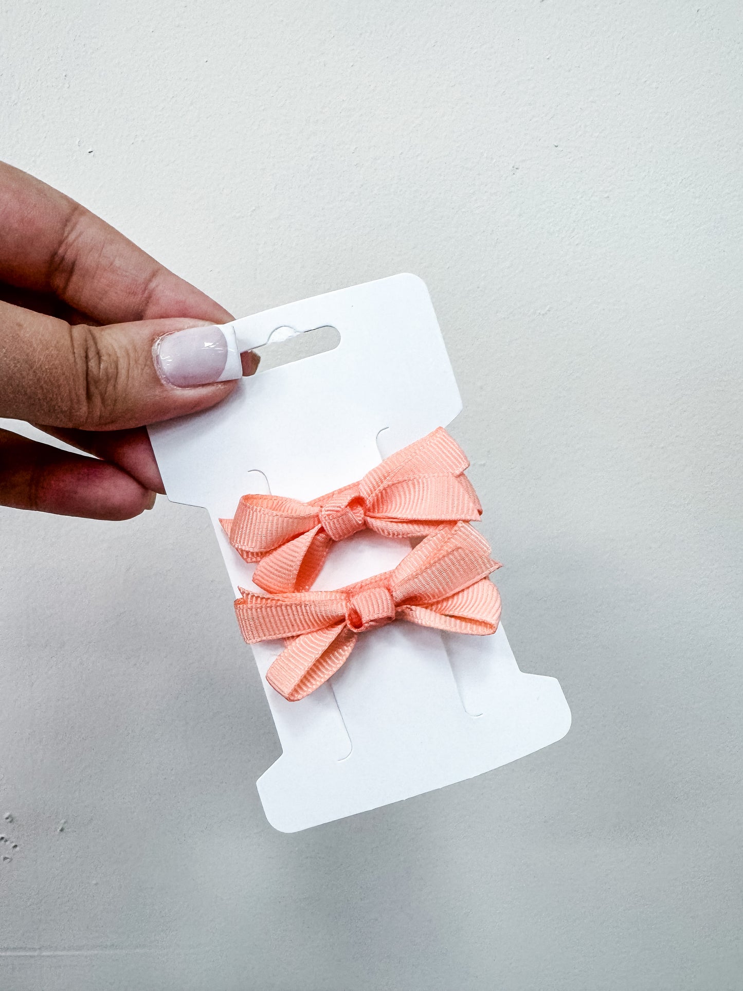 A hand holds a white card featuring two peach-colored ribbon bows from the Tiny Tot Bundles Duo Piggy Clip Pack, set against a simple light background.