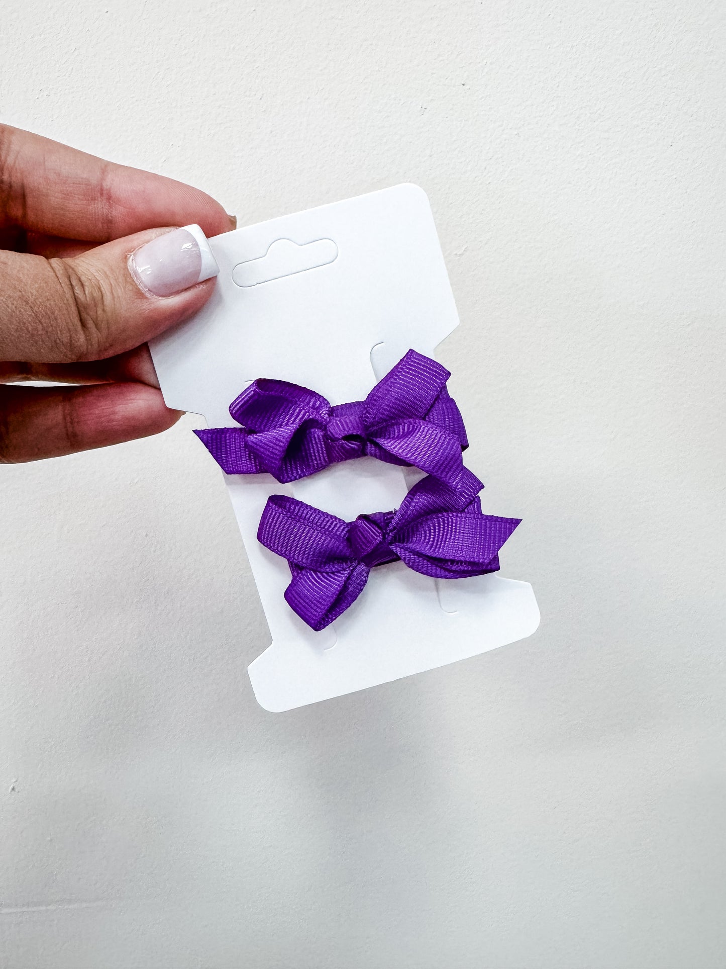 A hand holds a white card with two purple fabric bows from the Tiny Tot Bundles Duo Piggy Clip Pack, shown against a plain white background.