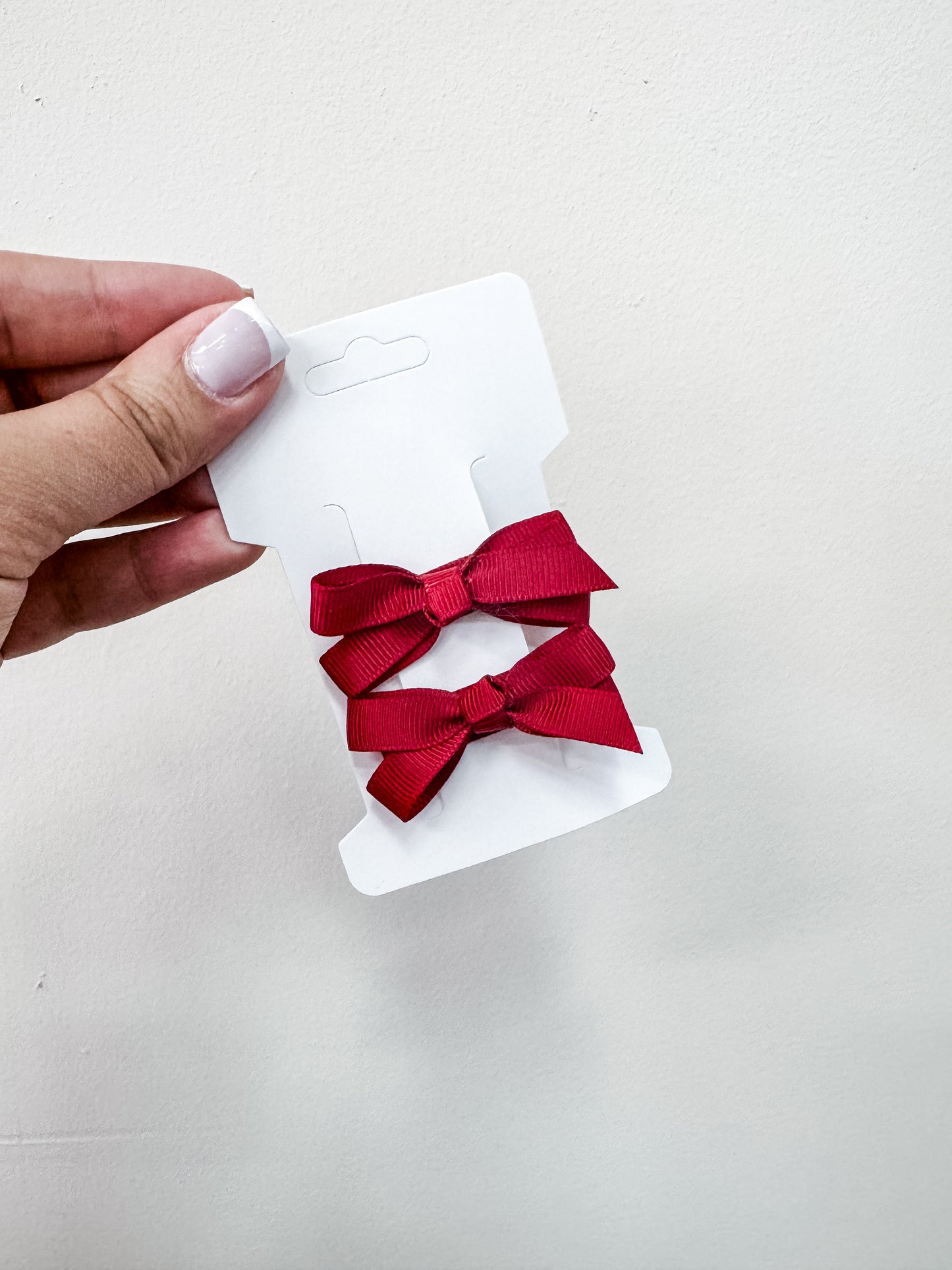 A hand with light nail polish holds a white card featuring two red ribbon bows from the Duo Piggy Clip Pack by Tiny Tot Bundles, set against a plain white background.