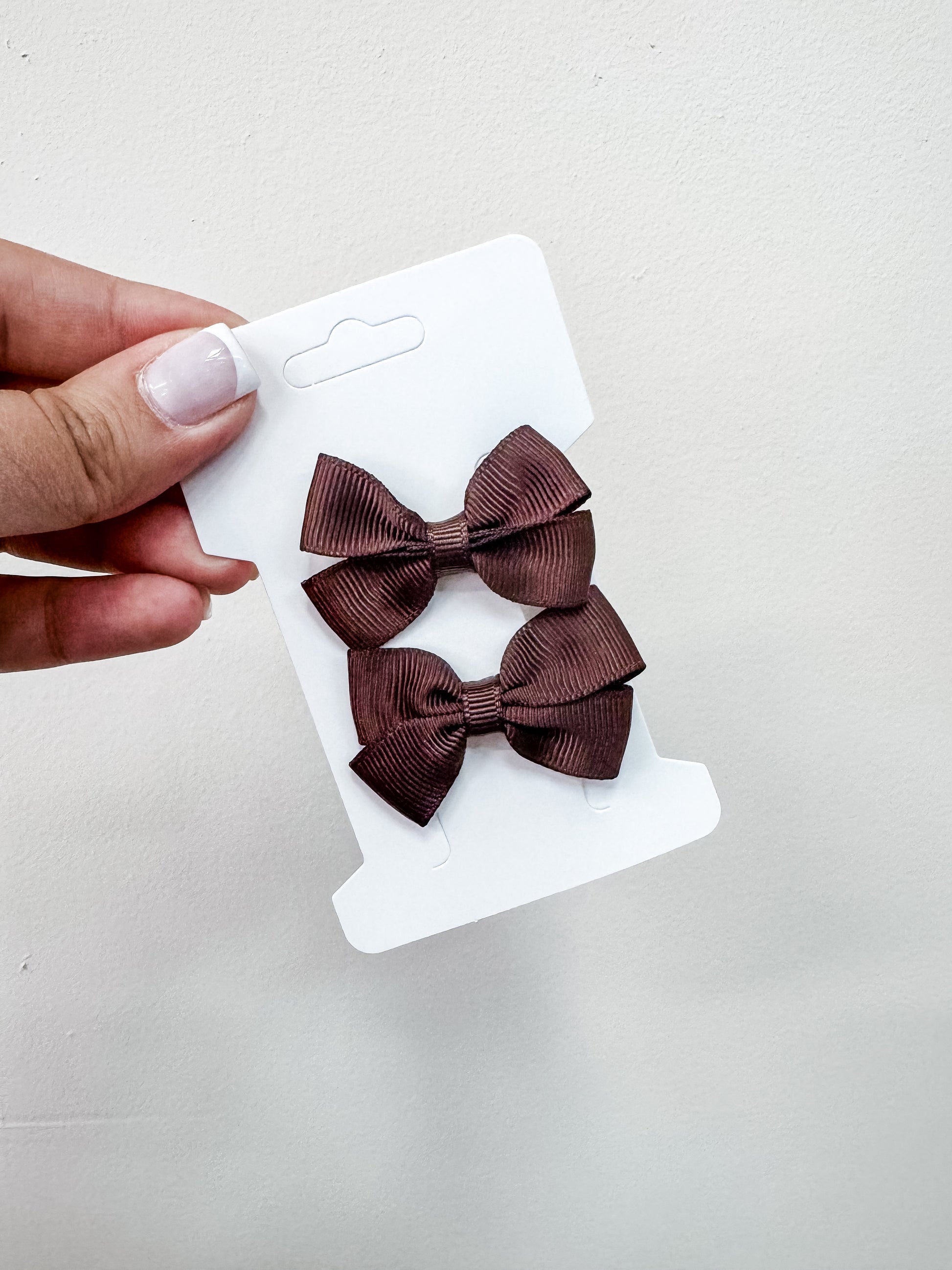 A hand with light pink nail polish holds a white card displaying the Tiny Tot Bundles Duo Piggy Clip Pack—two brown hair bows—against a plain, light background.