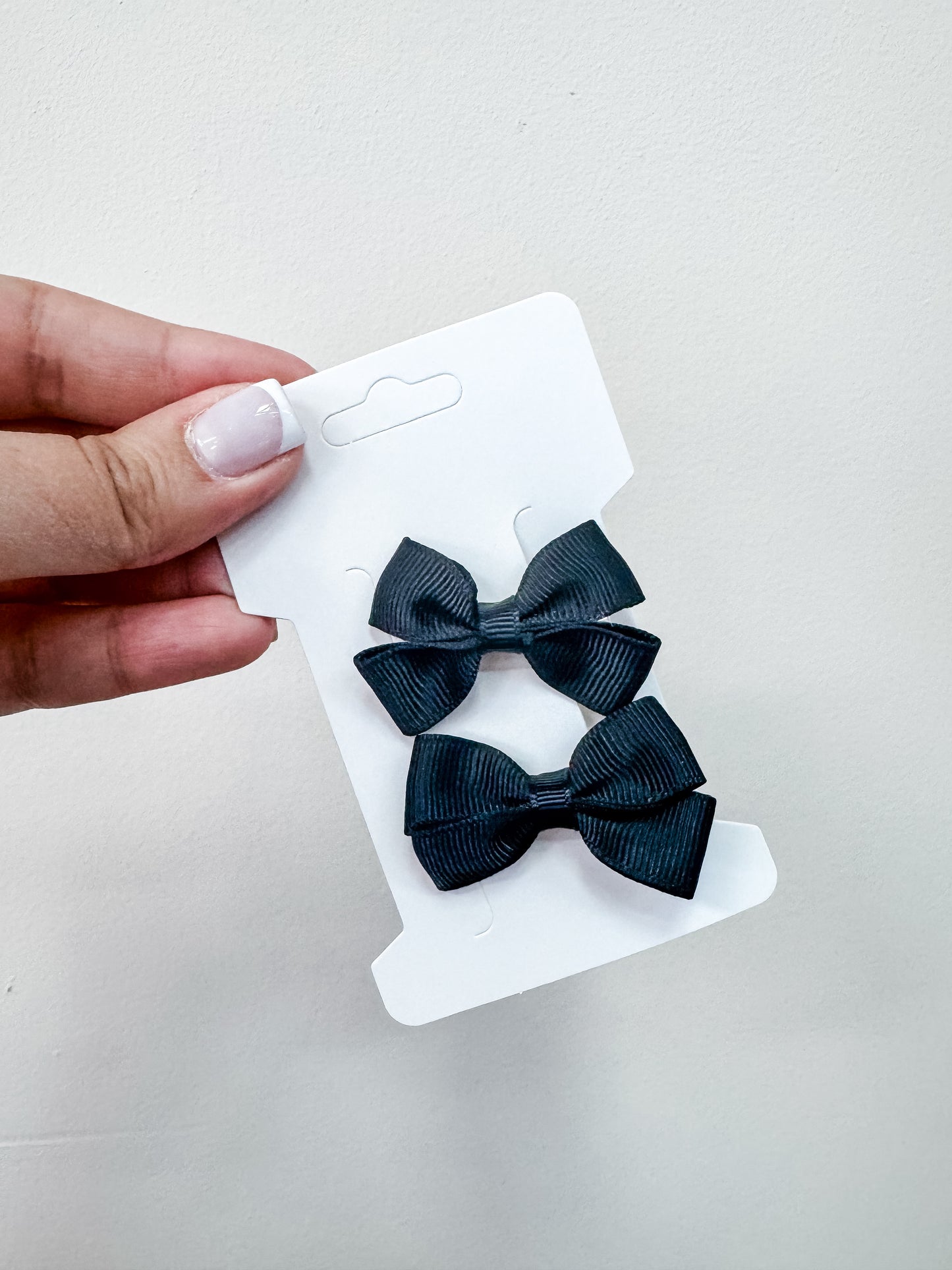 A hand with light-toned nails holds a white card showing two small, dark blue fabric bows from the Duo Piggy Clip Pack by Tiny Tot Bundles against a plain light background.