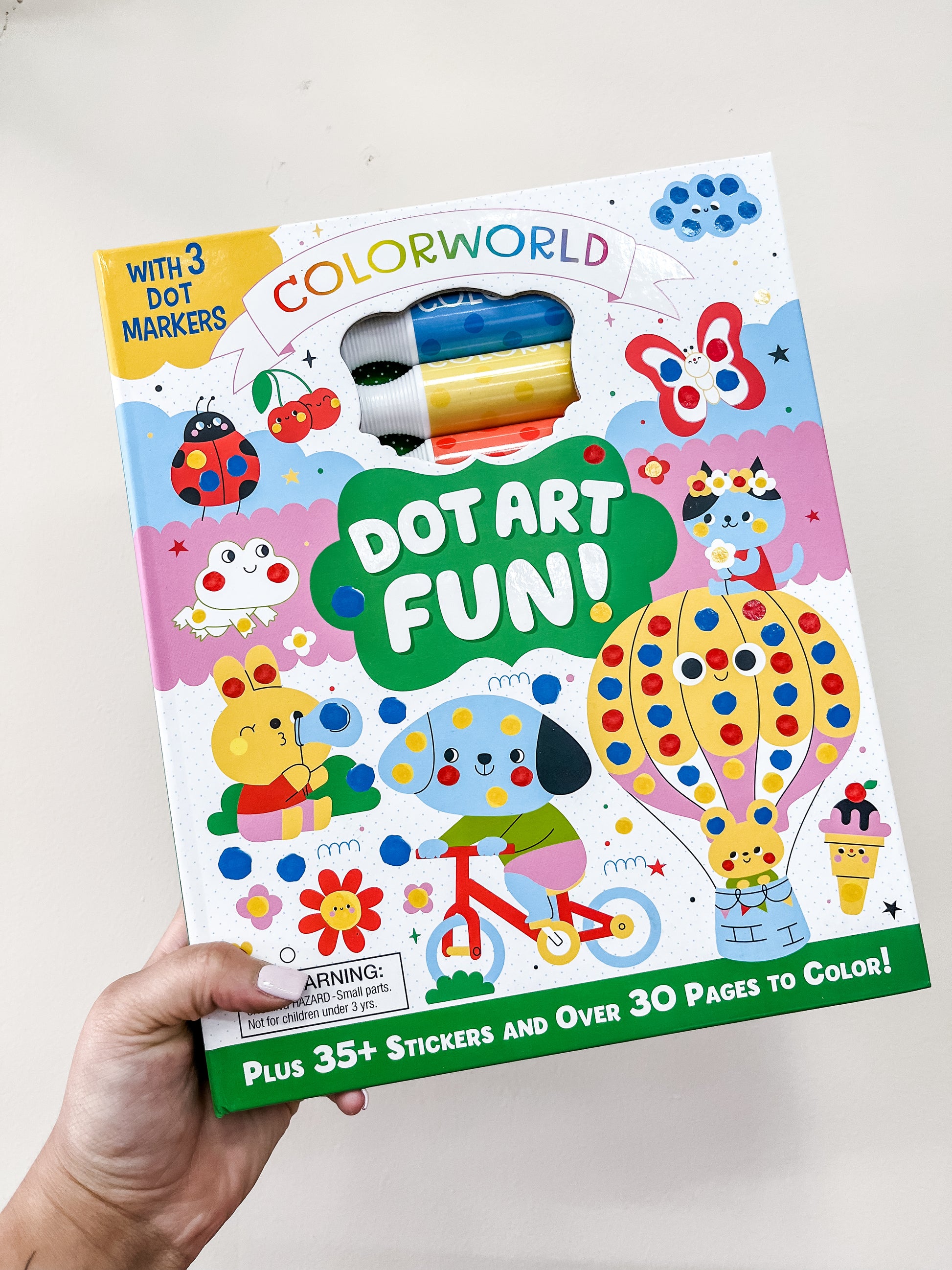 A hand holds the Simon & Schuster Color World - Dot Art Fun Activity Book with a colorful animal-themed cover. The set includes 3 dot markers, 35+ stickers, and over 30 coloring pages.