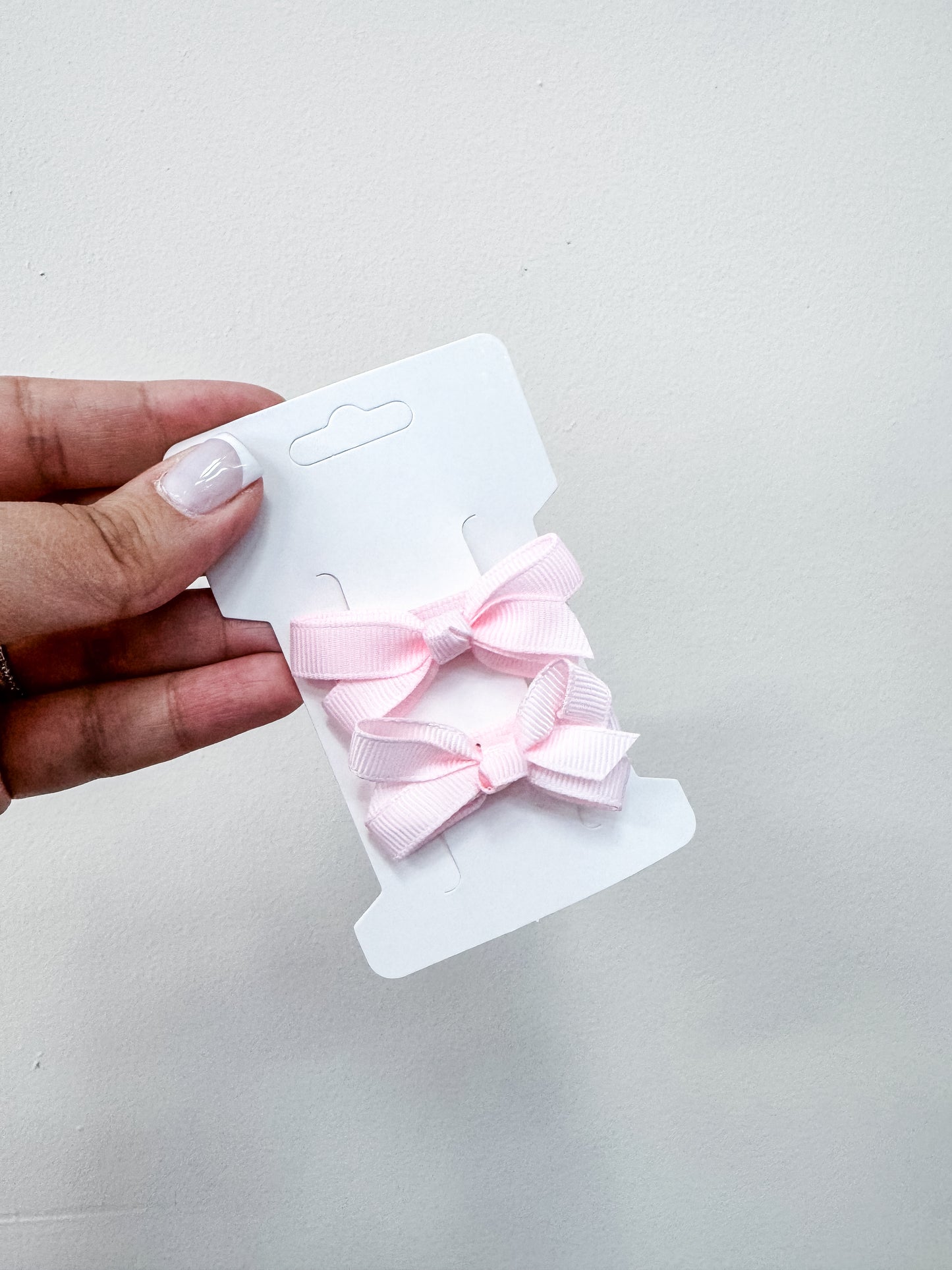 A hand with light pink nails holds a white card featuring two small, light pink Duo Piggy Clip Pack bows by Tiny Tot Bundles against a plain background.
