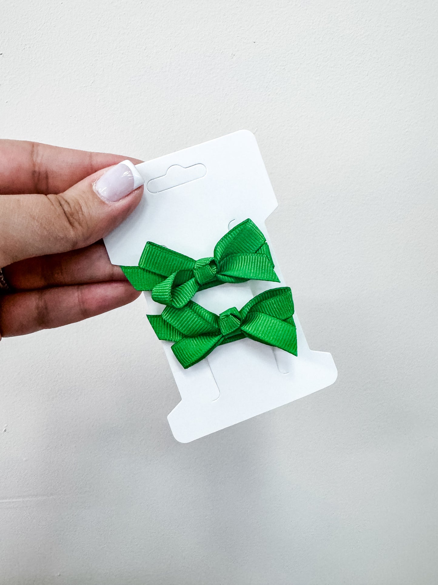A hand with a light pink manicure holds a white card displaying two bright green Duo Piggy Clip Pack bows by Tiny Tot Bundles against a plain white background.