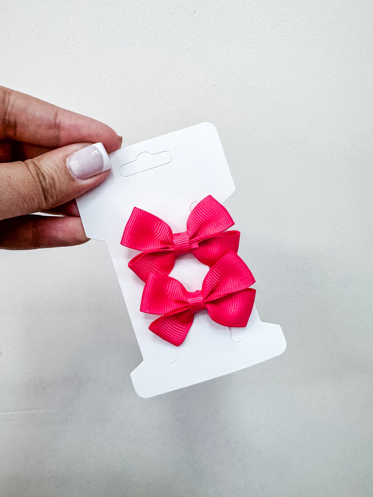A hand with manicured nails holds a white card featuring the Tiny Tot Bundles Duo Piggy Clip Pack—two bright pink hair bows—against a plain light background.