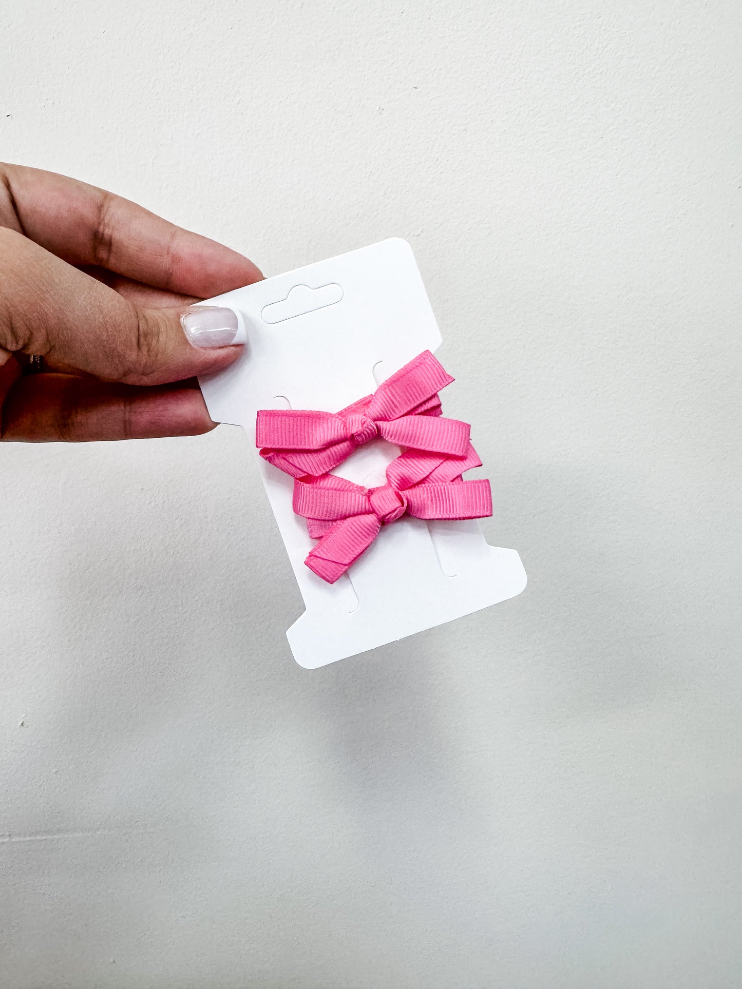 A hand with light nail polish holds a white card featuring the Duo Piggy Clip Pack—two bright pink ribbon bows by Tiny Tot Bundles—against an off-white background.