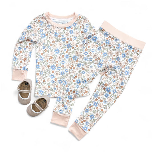 The Wildflower Whisper Jammies Set by Tiny Tot Bundles, featuring a pink, blue, and brown floral matching top and bottom, is displayed next to small beige shoes with white trim on a white background.