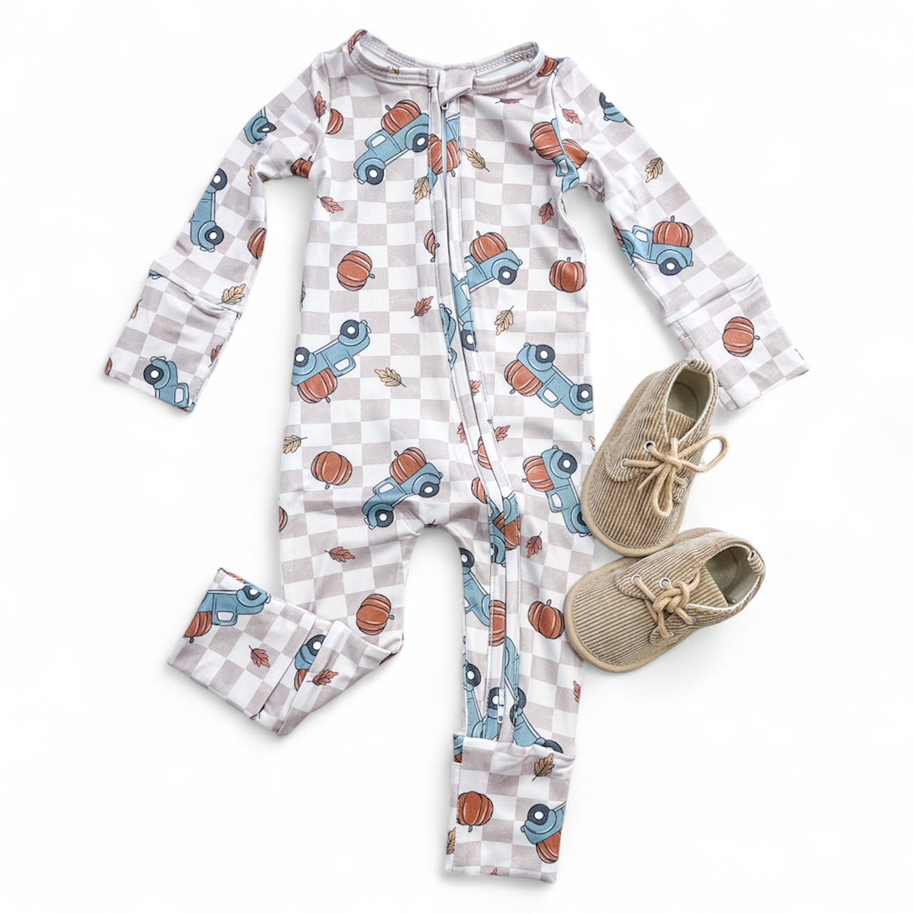 The Tiny Tot Bundles Fall Farm Run Zippy, a long-sleeve baby onesie with blue trucks, pumpkins, and leaves on a checkered background, is laid flat next to light brown baby shoes.