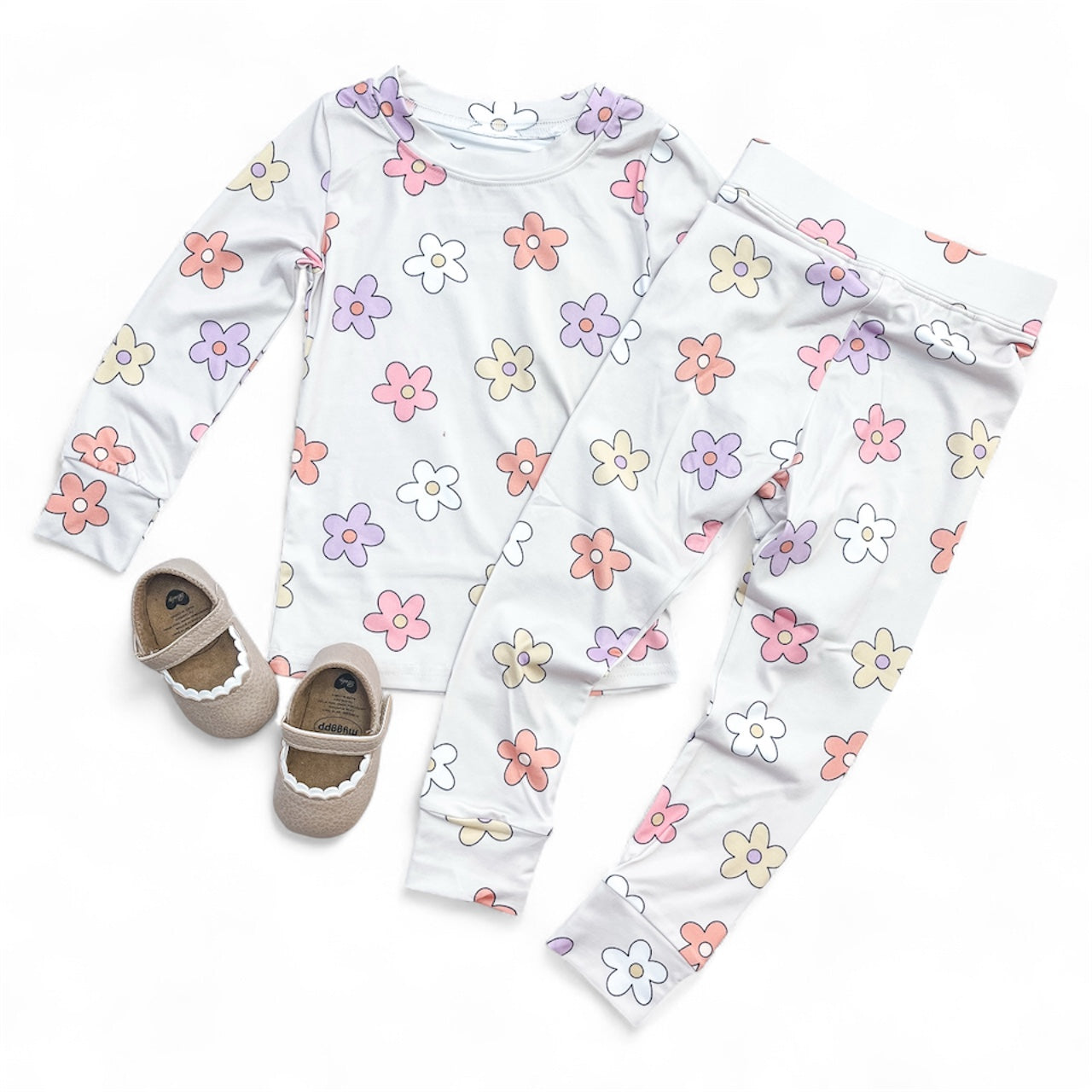 The Groovy Garden Jammies Set by Tiny Tot Bundles features long-sleeve pajamas with a pastel floral design in pink, purple, orange, and white. Light brown baby shoes with white scalloped trim are displayed beside the set.